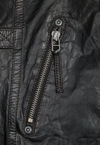 Dettaglio di una giacca di pelle nera con una zip argento prominente e cuciture marroni scure. La trama è leggermente spiegazzata e consumata.