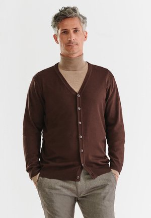 Brązowy kardigan z dekoltem w kształcie litery V, zapinany na guziki, z długimi rękawami. Noszony na beżowy golf. Wykonany z miękkiego, teksturowanego materiału dzianinowego.