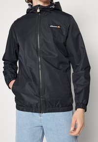 Czarna lekka kurtka z zamkiem błyskawicznym, elastycznymi mankietami i kapturem. Posiada teksturowane wykończenie oraz logo na piersi.