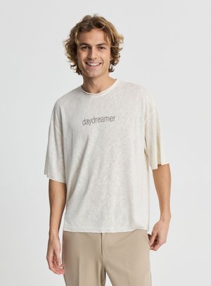 Giovane uomo con capelli ricci sorridente, che indossa una maglietta bianca larga con la scritta "daydreamer" e pantaloni beige su uno sfondo semplice.