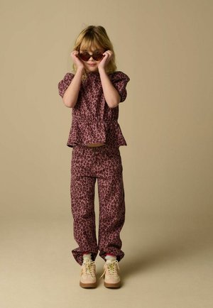 Girl wearing purple leopard print top and pants adjusts heart-shaped sunglasses, standing against plain beige background.