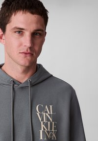 Felpa grigia con cappuccio caratterizzata da una superficie testurizzata, con cordini e un grande logo "CALVIN KLEIN" di colore chiaro sul fronte.
