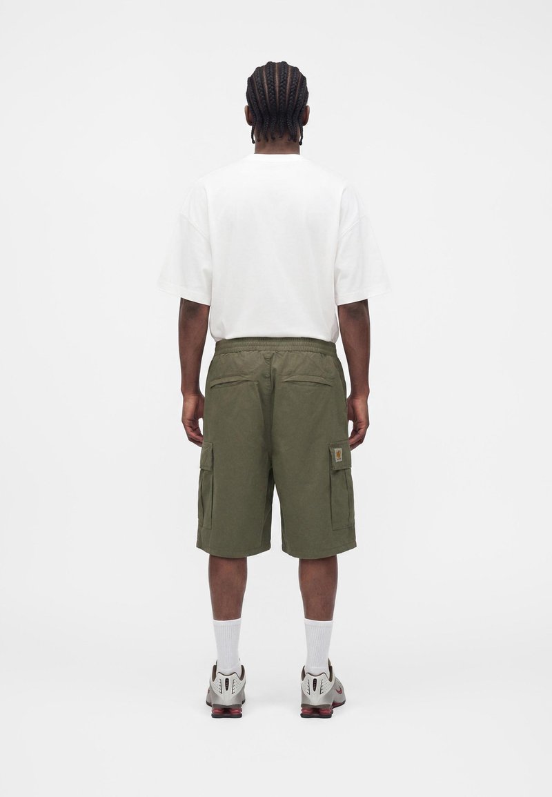 Mann mit geflochtenem Haar, der ein weißes übergroßes T-Shirt, olivgrüne Cargoshorts, weiße Socken und weiße Sneakers trägt, steht mit dem Rücken zur Kamera.