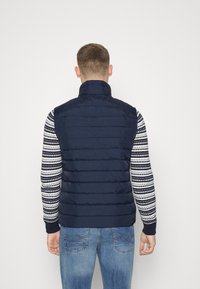 Marineblaue Steppweste mit hohem Kragen, ausgestattet mit horizontalem Steppmuster. Getragen über einem gemusterten Pullover und kombiniert mit hellblauen Jeans.