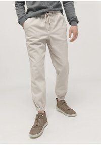 Light beige cotton jogger pants with elastic cuffs and adjustable drawstring waist. Paired with brown high-top sneakers.