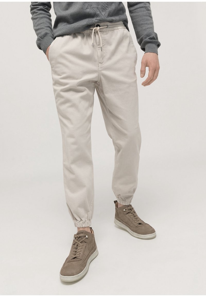 Light beige cotton jogger pants with elastic cuffs and adjustable drawstring waist. Paired with brown high-top sneakers.