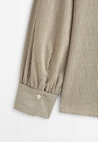 Chemise à manches longues rayée en beige et noir, avec manches froncées, poignets boutonnés et tissu texturé à fines lignes verticales.