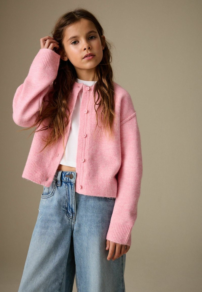 Cardigan corto rosa chiaro realizzato in morbido tessuto a maglia, con chiusura frontale con bottoni, polsini a costine e vestibilità morbida, abbinato a jeans di denim azzurro chiaro.