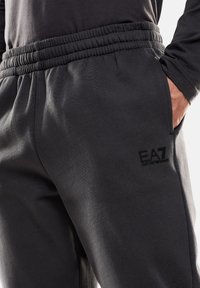 Pantaloni sportivi grigio scuro con vita elasticizzata, caratterizzati da un logo EA7 nero. Il materiale sembra liscio e presenta una texture morbida. Sono presenti tasche.