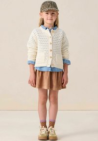 Strickcardigan in Creme mit Rundhalsausschnitt und goldenen Knöpfen, über einem blauen Hemd getragen, kombiniert mit einem braunen Cordrock und beigen Sneakers.