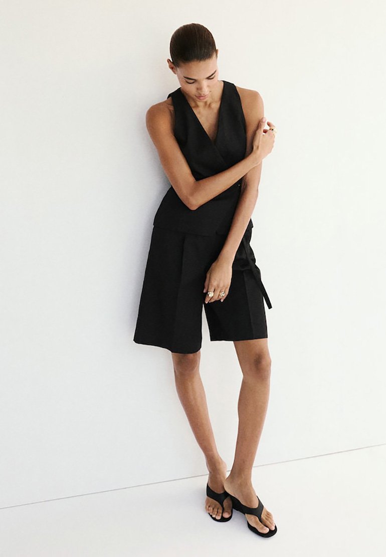 Femme portant un haut noir sans manches et un short noir longueur genoux, debout contre un mur blanc, les bras croisés.