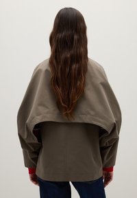 Khaki oversized jacket with a loose fit, featuring a wide back flap and red accents on the sleeves, made of a smooth fabric.