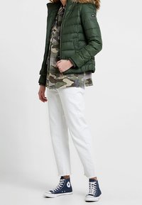 Grön puffjacka med foder i päls, kamouflageknäppt skjorta, vita cropped byxor och blå höga sneakers.