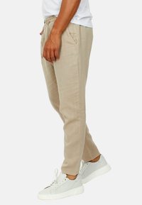 Pantaloni in lino beige con vestibilità rilassata, vita elastica e tasche laterali, abbinati a sneaker da ginnastica grigio chiaro.