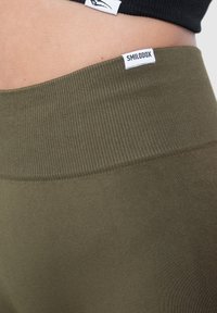 Leggings sportivi color verde oliva con un alto cinturino a costine. Presenta un'etichetta bianca con il marchio "SMILODOX". Texture liscia.