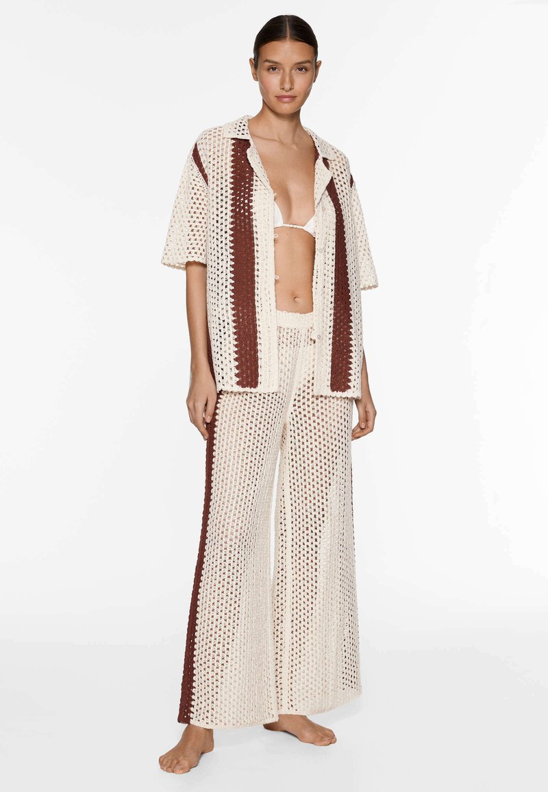 Donna in piedi a piedi nudi, che indossa una camicia aperta in pizzo color crema e marrone, pantaloni larghi abbinati e un bikini bianco.