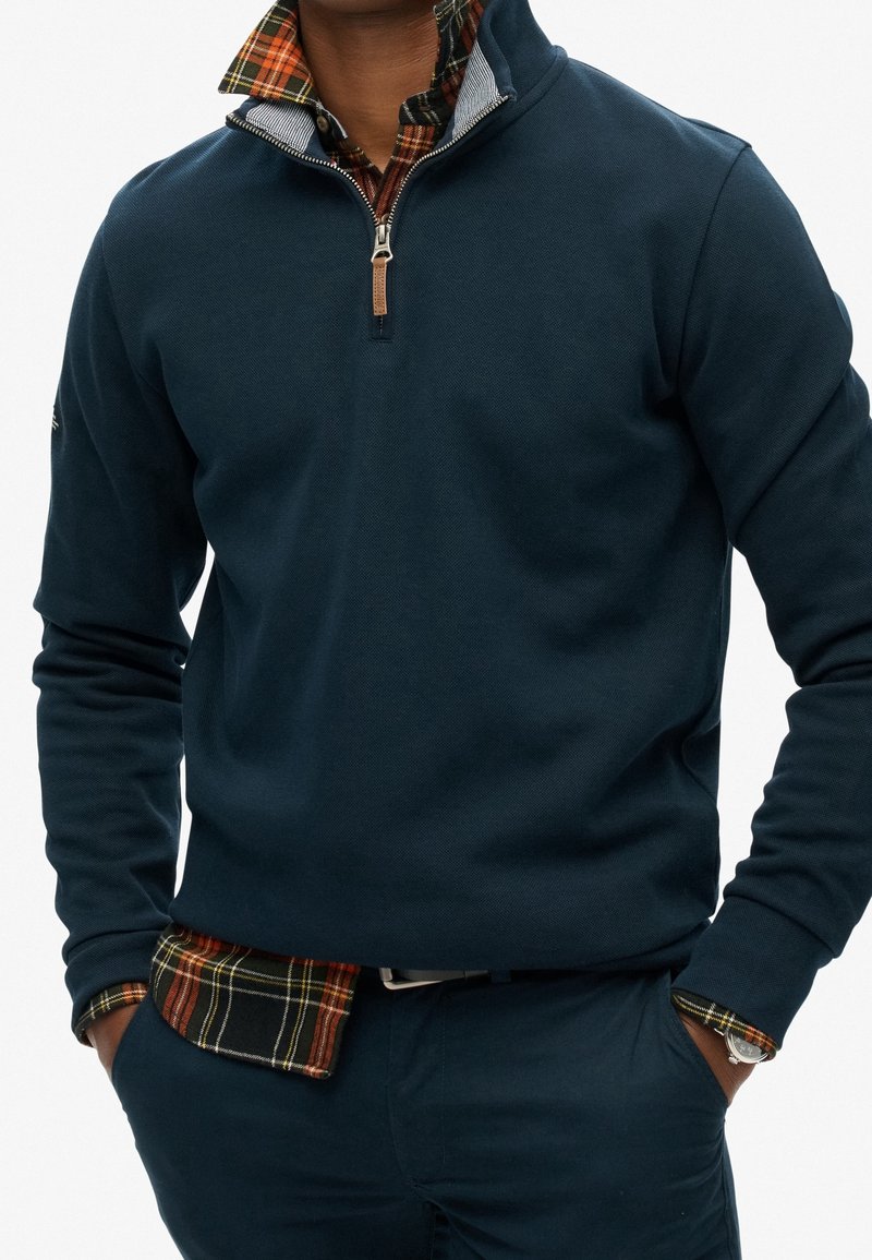 Pull-over bleu marine avec col à fermeture éclair partielle, superposé à une chemise à carreaux aux tons orange et vert, tissu texturé.