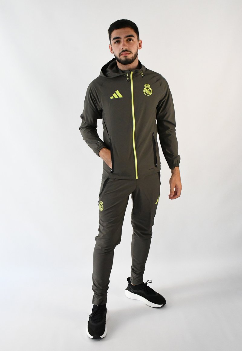 Chándal atlético gris con acentos amarillos, que incluye una chaqueta con cremallera y pantalones ajustados. Presenta el logo del Real Madrid y las franjas de Adidas. Zapatillas negras.