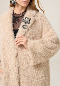 Beige, načechraný kabát ve velkém střihu s texturovaným povrchem. Obsahuje květinovou brož se stříbrnými akcenty. Nosený přes černý krajkový top.