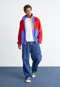 Giacca in pile rossa e viola con dettagli arancioni, chiusura con zip, colletto alto; abbinata a pantaloni cargo blu larghi e scarpe da ginnastica.
