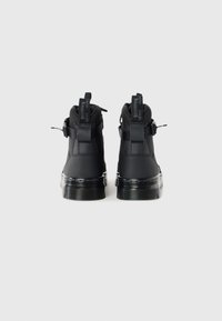 Bottes montantes noires avec semelles épaisses translucides, languettes arrière avec texte et brides latérales, vues de dos sur un fond uni.