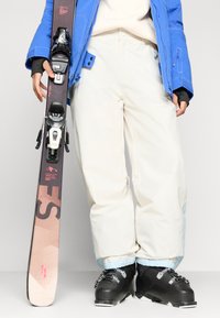 Esqui com design gráfico em preto e rosa, segurado por uma pessoa em calças de neve brancas e uma jaqueta azul com punhos e fechos em contraste.