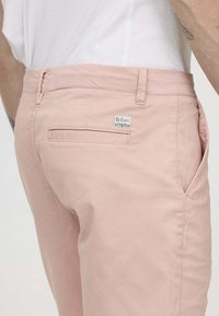 Pantalon en coton rose clair avec une coupe slim, dotée d'une poche à l'arrière et d'une étiquette sur la ceinture. Texture lisse, détails de couture minimalistes.