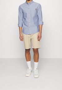 Camicia azzurra chiara a maniche lunghe con taschino sul petto, abbinata a pantaloni beige e calzini bianchi. Le scarpe sono sneakers grigio chiaro con dettagli beige.