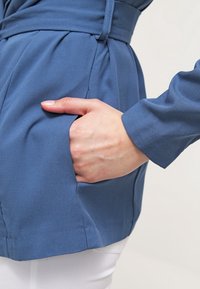 Main reposant dans la poche d'une veste bleue ceinturée portée sur un pantalon blanc, sur un fond neutre.