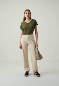 Mujer joven con una blusa de manga corta verde oliva, pantalones de cintura alta beige, zapatillas negras, sosteniendo un bolso beige, de pie contra un fondo claro y sencillo.