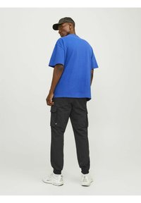 Blå oversized crewneck t-shirt, sorte cargo bukser med sidelommer og hvide sneakers. Model bærer en sort cap, set fra bagfra.