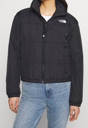 Person iført en sort North Face lynlåspufferjakke med høj krave og lyseblå jeans, stående foran en ensfarvet baggrund.