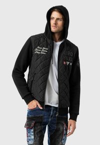 Schwarze gesteppte Jacke mit Kapuze, die mit gesticktem Text und Patches versehen ist, kombiniert mit blauen Jeans-Cargohosen mit mehreren Taschen.