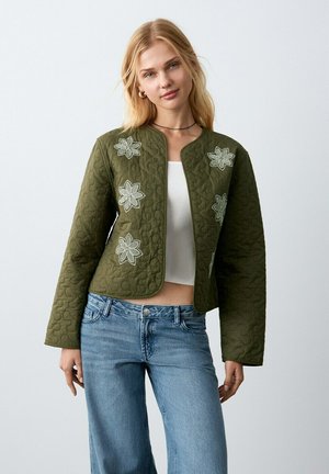 Donna bionda che indossa una giacca trapuntata verde oliva con fiori ricamati, top corto bianco e jeans blu, in piedi davanti a uno sfondo semplice.