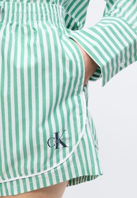 Persona valkā zaļi baltas vertikāli svītrainas šortus un atbilstošu kreklu, roka ielikta šortu kabatā, redzams Calvin Klein logotips.