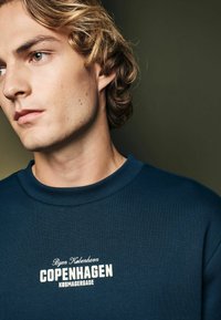 Námornícky sveter s rebrovaným kulatým výstrihom, s bielym textom "Byen København COPENHAGEN KØBMAGERGADE" umiestneným na strede hrudníka.