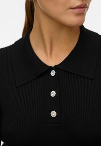 Mustmustriibuline polo särk mustas toonis, millel on kaeluses kolm dekoratiivset hõbedast lillekujulist nööpi. Sile tekstuur ja struktureeritud disain.
