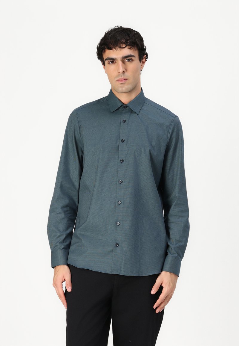 Chemise boutonnée en bleu sarcelle foncé en tissu texturé, avec un col pointu, des manches longues et des boutons noirs. Portée avec un pantalon noir.