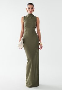 Robe maxi en maille côtelée vert olive avec col montant et silhouette ajustée. Accessoirisée d'un sac à main crème et de boucles d'oreilles créoles dorées. Texture lisse.