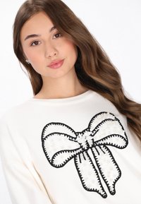 Biała sweter z dużym czarno-białym wzorem w kształcie kokardy, z teksturalnymi detalami i ściegami. Gładki materiał, luźny krój.