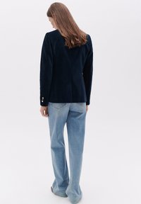 Marineblauwe suède blazer met gestructureerde schouders, enkele rugventilatie en gouden knopen, gecombineerd met lichtblauwe wijde jeans.