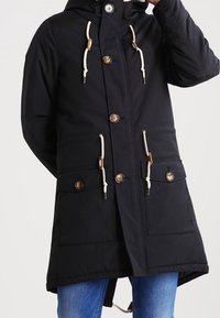 Svart parka jacka med huva, knappknäppning, två framfickor och dragsnören. Tillverkad av texturerat tyg.