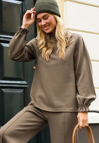 Vrouw met een olijfgroen gebreid mutsje en bijpassende losse top en broek, die een beige handtas vasthoudt, glimlacht voor een donkergrode deur.