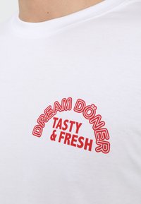 Bjela pamučna majica s crvenim grafičkim dizajnom koji prikazuje natpis "DREAM DÖNER TASTY & FRESH" u podebljanim, zakrivljenim slovima.