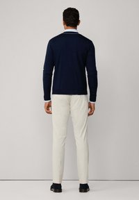 Uomo con capelli ricci scuri che indossa un maglione blu navy, una camicia azzurro chiaro, pantaloni color crema e scarpe nere, in piedi di fronte a un muro semplice.