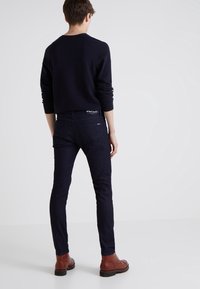 Marinblå ribbad stickad tröja med långa ärmar, kombinerad med mörka jeans och bruna läderskor. Visad bakifrån mot en ljus bakgrund.