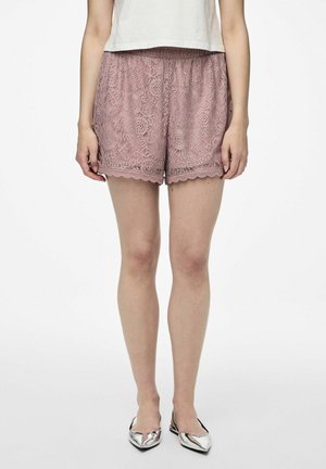 Kvinde iført lave shorts i mauve blonde, hvid top og metallisk sølvfarvede spidse flade sko, stående mod en hvid baggrund.