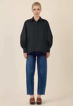 Blonde woman stands wearing loose black button-up shirt, relaxed blue jeans, and brown platform sandals against plain beige background.