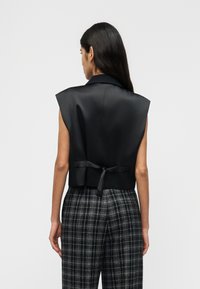 Γυναίκα με μακριά σκούρα μαλλιά, φορώντας ασπρόμαυρο καρό παντελόνι και αμάνικο μαύρο σατέν γιλέκο δεμένο στο πίσω μέρος, στέκεται μπροστά σε λευκό φόντο.