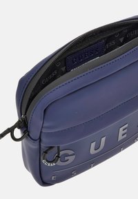 Bolso sintético azul marino con el logotipo "GUESS" en relieve, cierre de cremallera y cordón negro ajustable. Presenta un forro interior con un patrón.
